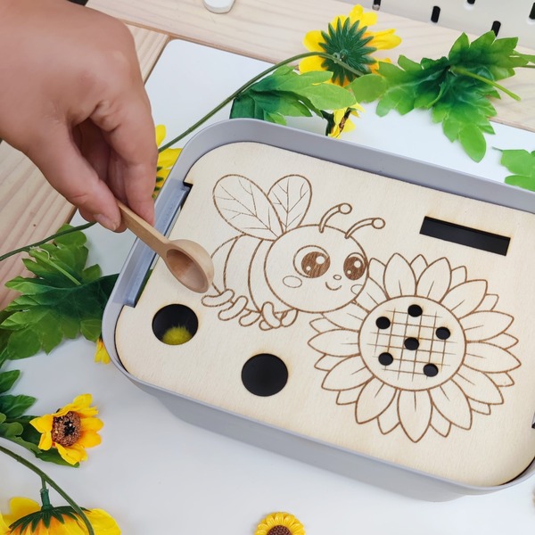 Enfant utilisant une cuillère en bois pour déposer un pompon dans un insert abeille et tournesol en bois