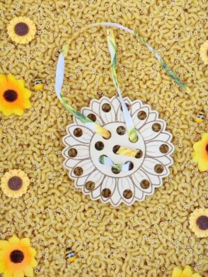 Carte à lacer en bois en forme de tournesol avec ruban coloré, posée sur un bac sensoriel décoré de fleurs et pâtes sèches