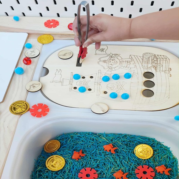Enfant utilisant une pince sur un insert pirate en bois avec jetons et trésors sensoriels