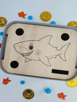 Insert en bois motif requin posé dans un bac sensoriel avec pièces dorées, poissons orange et pompons bleus