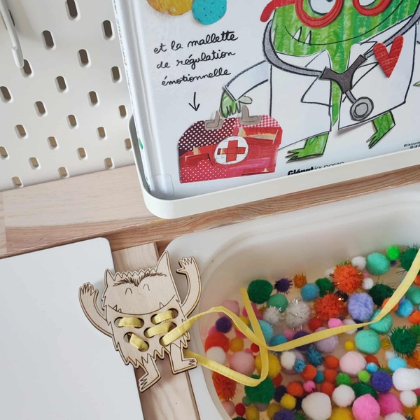 Carte à lacer en bois en forme de monstre utilisée avec un lacet jaune et des pompons sensoriels pour stimuler la motricité fine des enfants