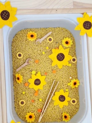 Bac sensoriel rempli de pâtes avec fleurs en feutrine tournesol, pompons colorés et petits outils en bois