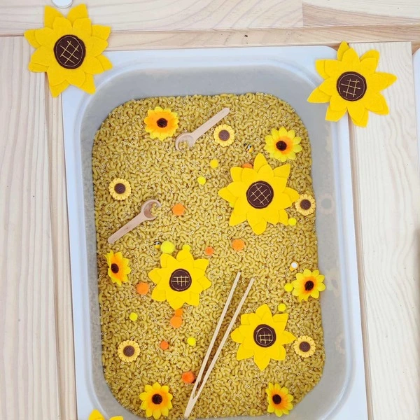 Bac sensoriel rempli de pâtes avec fleurs en feutrine tournesol, pompons colorés et petits outils en bois