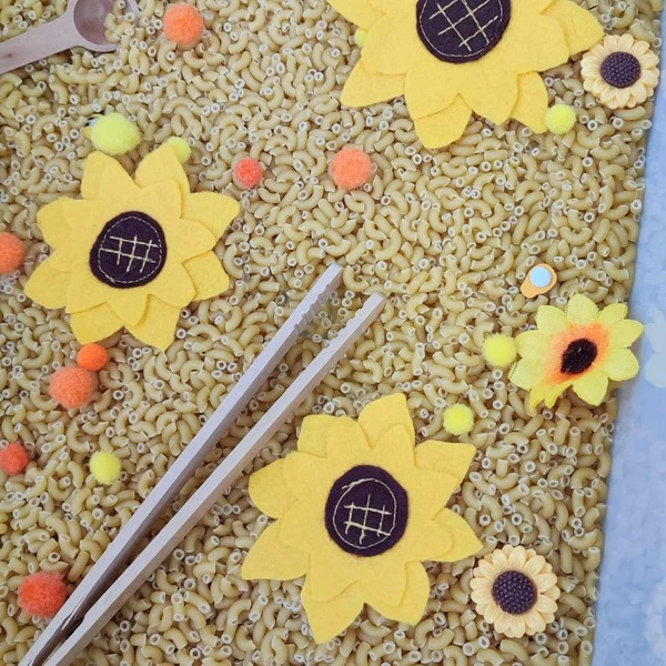 Gros plan sur un bac sensoriel avec pâtes, pompons colorés, tournesols en feutrine et baguettes en bois