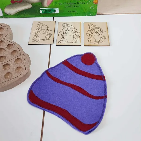 Bonnet en feutrine violette et rouge accompagné de cartes en bois gravées représentant le lapin du livre “Le Bonnet de Lapinus”