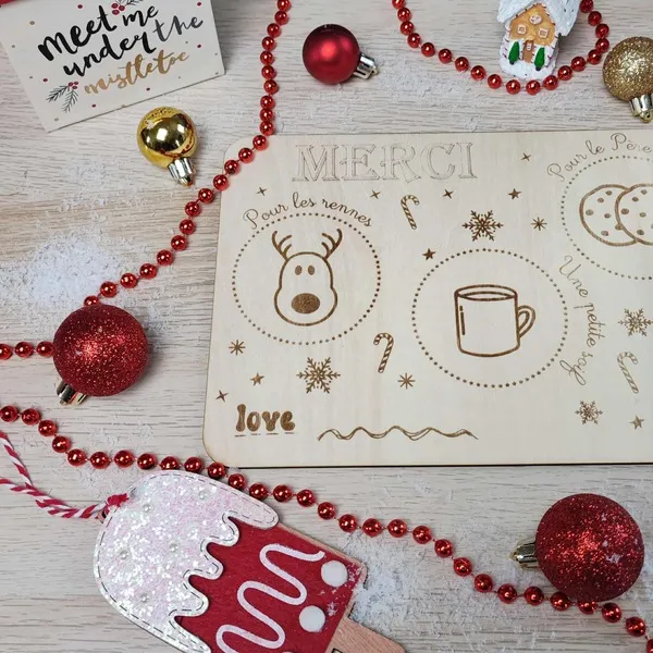Détail d’un plateau en bois gravé “Merci Père Noël” entouré de boules rouges, guirlande et décorations de Noël artisanales
