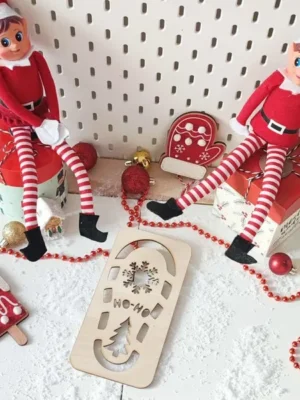 Mise en scène de Noël avec deux lutins farceurs et une empreinte en bois “Ho Ho Ho” entourée de neige artificielle et décorations rouges