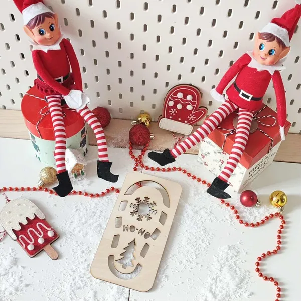 Mise en scène de Noël avec deux lutins farceurs et une empreinte en bois “Ho Ho Ho” entourée de neige artificielle et décorations rouges