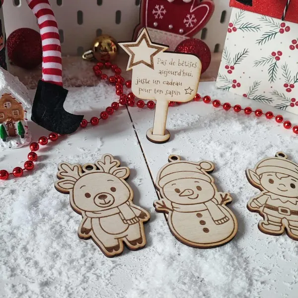 Décor de Noël enneigé avec ornements en bois gravé représentant un renne, un bonhomme de neige, un lutin et un panneau message “Pas de bêtises aujourd’hui, juste un cadeau pour ton sapin”