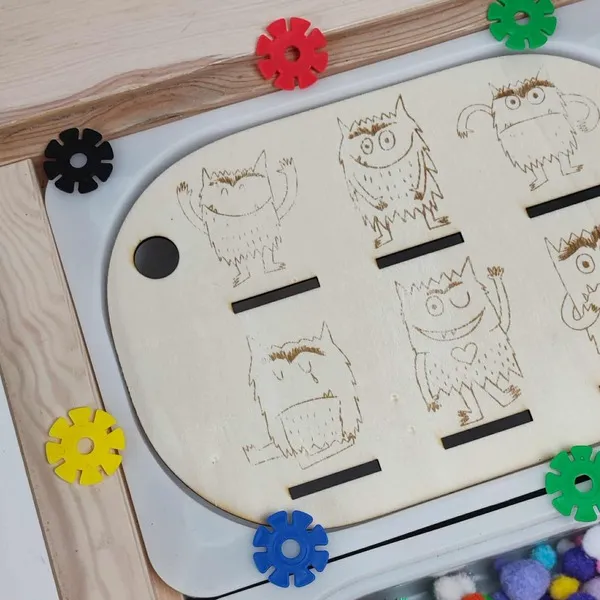 Insert en bois gravé représentant les monstres des émotions, installé sur un bac sensoriel avec pompons et disques colorés