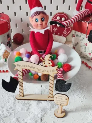 Lutin farceur de Noël installé dans une piscine à balles colorées avec pancartes en bois gravé “Piscine à balles réservée au lutin” et “Risque d’éclaboussures de bêtises”