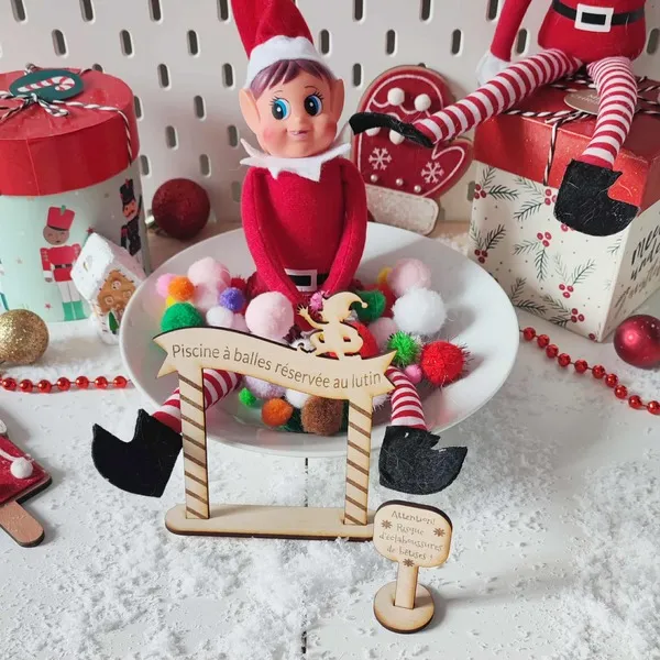 Lutin farceur de Noël installé dans une piscine à balles colorées avec pancartes en bois gravé “Piscine à balles réservée au lutin” et “Risque d’éclaboussures de bêtises”