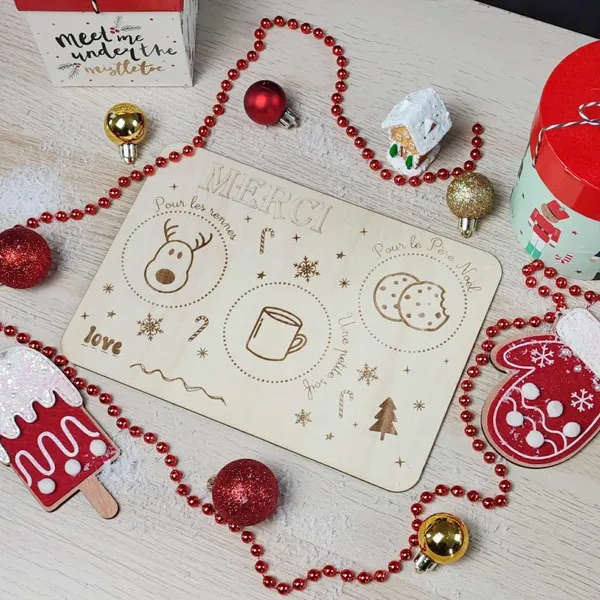 Plateau en bois gravé “Merci Père Noël” entouré de décorations de Noël rouges et dorées, avec guirlande, boules et petites boîtes cadeaux
