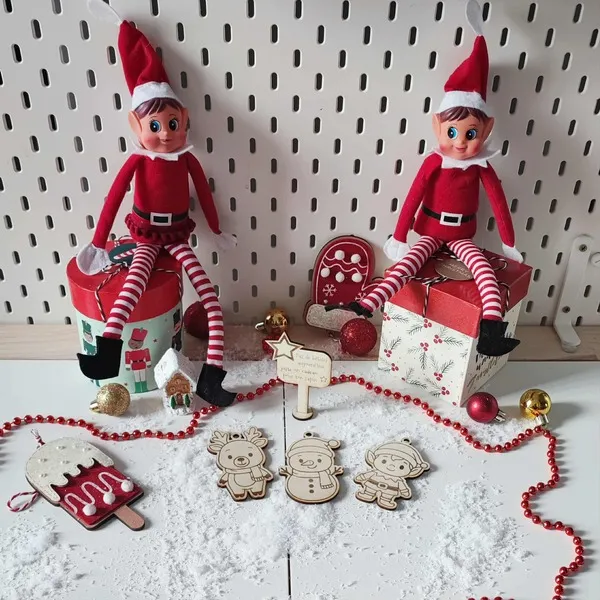 Décor de Noël avec deux lutins farceurs et des ornements en bois gravé représentant un renne, un bonhomme de neige et un lutin, entourés de neige artificielle et de guirlandes rouges