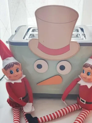 Deux lutins farceurs de Noël assis devant un grille-pain transformé en bonhomme de neige avec chapeau, yeux et nez en bois coloré