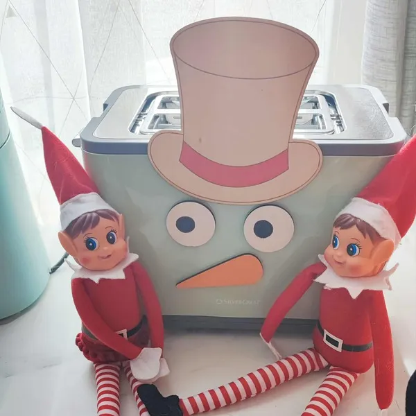 Deux lutins farceurs de Noël assis devant un grille-pain transformé en bonhomme de neige avec chapeau, yeux et nez en bois coloré