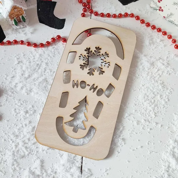 Empreinte en bois “Ho Ho Ho” avec flocon et sapin posée sur de la fausse neige, décor de Noël avec guirlande rouge et maison pain d’épices