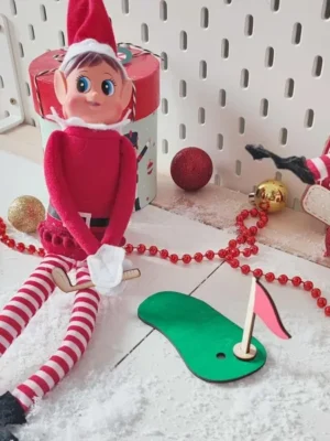 Lutin farceur assis dans un décor de Noël enneigé, jouant au mini-golf avec accessoires en bois et drapeau rouge
