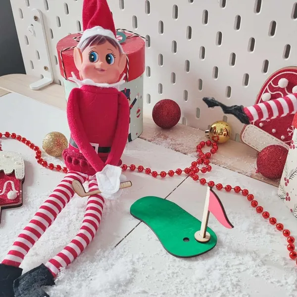 Lutin farceur assis dans un décor de Noël enneigé, jouant au mini-golf avec accessoires en bois et drapeau rouge