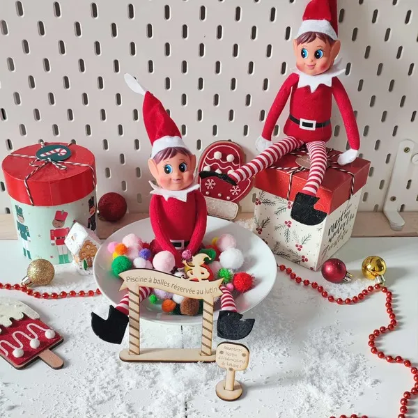 Deux lutins farceurs de Noël dans un décor enneigé, dont un assis dans une piscine à balles colorées avec pancartes en bois gravé “Piscine à balles réservée au lutin” et “Risque d’éclaboussures de bêtises”