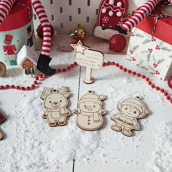 Décor de Noël enneigé avec trois ornements en bois gravé – renne, bonhomme de neige, lutin – et un panneau étoilé “Pas de bêtises aujourd’hui, juste un cadeau pour ton sapin”
