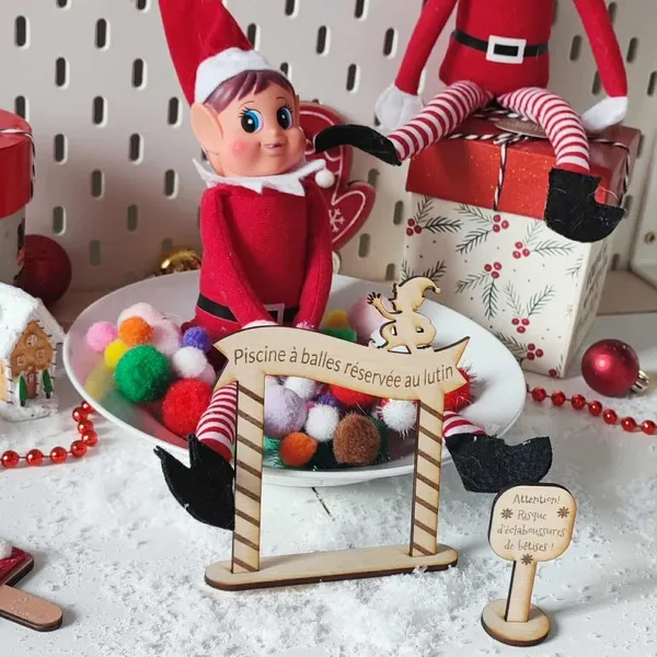 Lutin farceur de Noël assis dans une piscine à balles colorées avec panneaux en bois gravé “Piscine à balles réservée au lutin” et “Risque d’éclaboussures de bêtises”