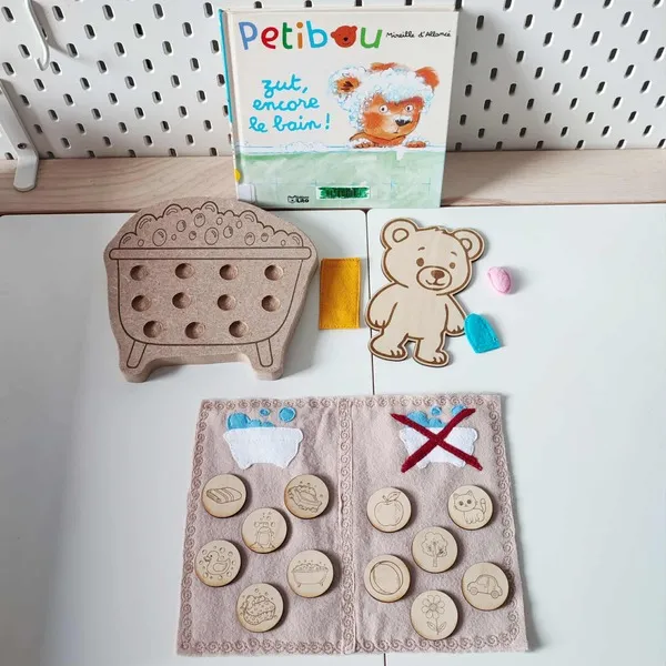 Activité éducative inspirée du livre “Petibou zut encore le bain !” avec ours en bois, baignoire à picots et jetons gravés sur le thème de l’hygiène et du quotidien