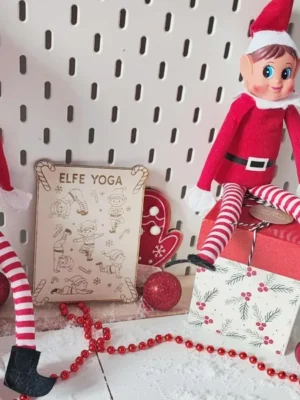 Plaque en bois gravée “Elfe Yoga” entourée de deux lutins farceurs dans un décor de Noël enneigé avec guirlandes et cadeaux
