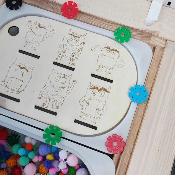 Plaque en bois gravée des monstres des émotions posée sur une table sensorielle avec pompons colorés
