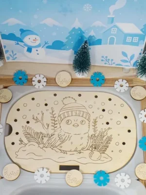 Plateau sensoriel en bois gravé d’un petit oiseau hivernal avec bonnet, décoré de jetons flocons et de sapins miniatures pour activité Montessori d’hiver