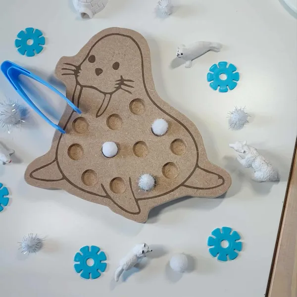 Plateau de motricité fine en bois en forme de morse, utilisé avec une pince bleue et des pompons blancs dans une activité Montessori sur le thème polaire