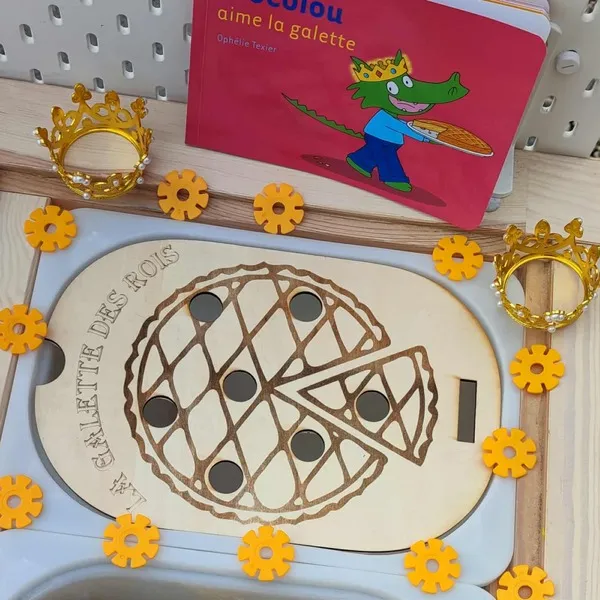 Plateau en bois gravé représentant une galette des rois avec jetons noirs, couronnes dorées et livre Crocolou aime la galette