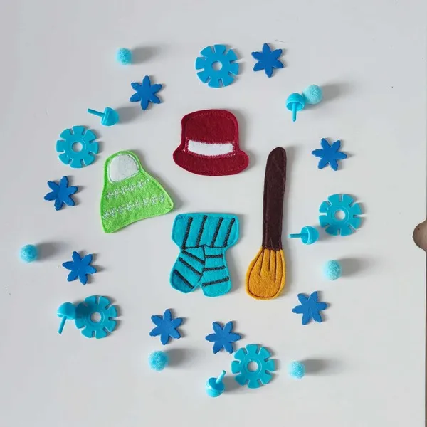 Accessoires en feutrine colorée pour habiller un bonhomme de neige, entourés de flocons et pompons bleus pour activité Montessori d’hiver