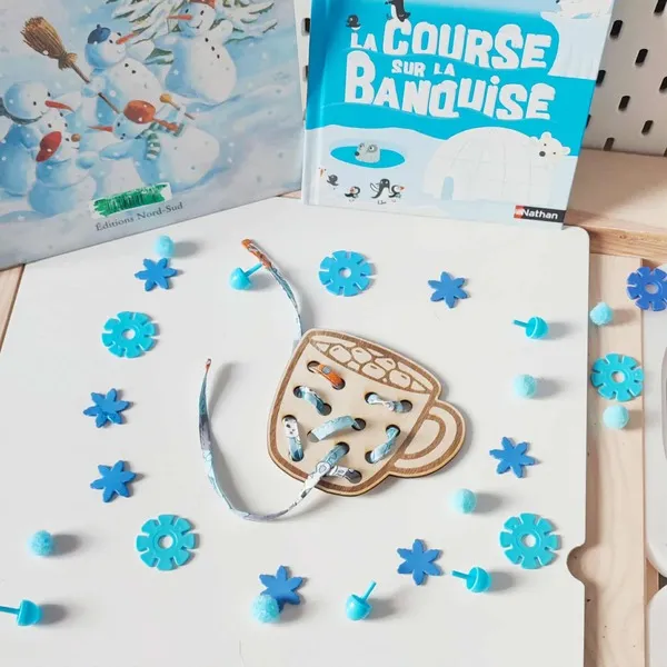 Carte à lacets en bois en forme de tasse de chocolat chaud, décorée sur le thème de l’hiver avec accessoires bleus et livres sur la banquise en arrière-plan
