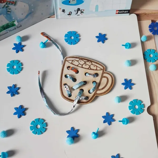 Carte à lacets en bois en forme de tasse, partiellement enfilée avec un ruban coloré, entourée d’éléments bleus sur le thème de l’hiver et de la motricité fine Montessori