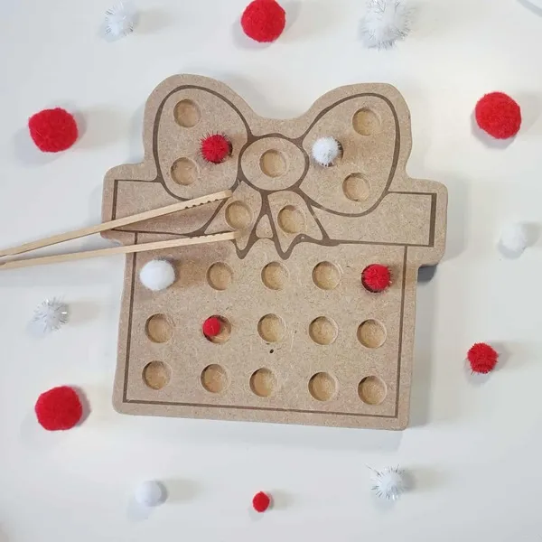 Plateau de motricité fine en bois MDF en forme de cadeau avec nœud, utilisé avec des pompons rouges et blancs et des baguettes sur fond clair