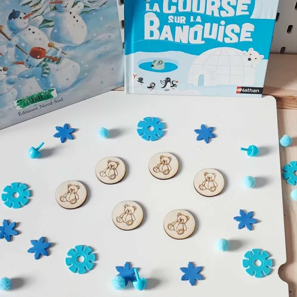 Jetons en bois gravés d’un ourson disposés sur une table décorée d’éléments bleus hivernaux, devant des livres sur la banquise et la neige