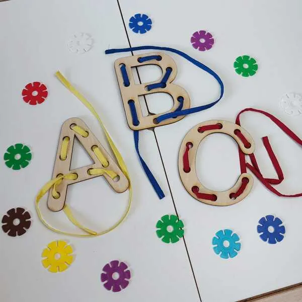 Lettres en bois à lacer posées sur fond blanc, entourées de petites formes colorées.