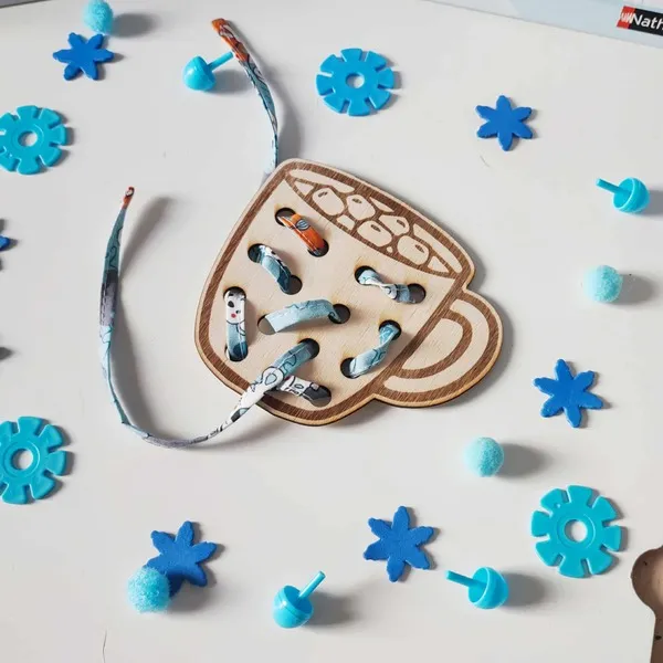 Carte à lacer en bois en forme de tasse avec ruban coloré, activité de motricité fine Montessori entourée d’éléments bleus évoquant l’hiver