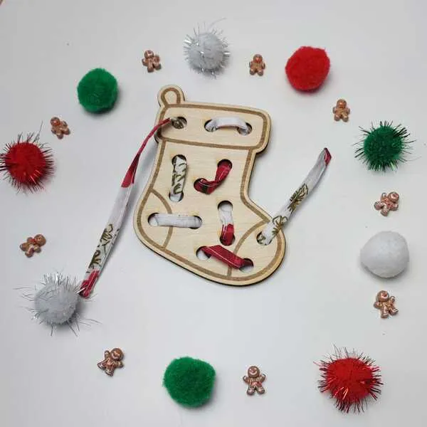 Carte à lacer en bois en forme de chaussette de Noël entourée de pompons colorés et de petites décorations festives.