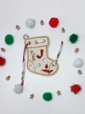 Carte à lacer en forme de chaussette de Noël, entourée de pompons colorés et de petites décorations.