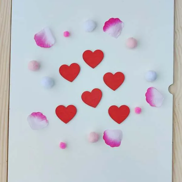 Cœurs rouges en bois disposés sur fond blanc avec pétales et pompons roses pour activité sensorielle enfant