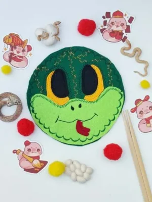 Masque serpent en feutrine verte entouré d’éléments sensoriels, pompons rouges et jaunes, thème Nouvel An chinois