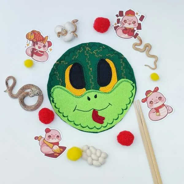 Masque serpent en feutrine verte entouré d’éléments sensoriels, pompons rouges et jaunes, thème Nouvel An chinois