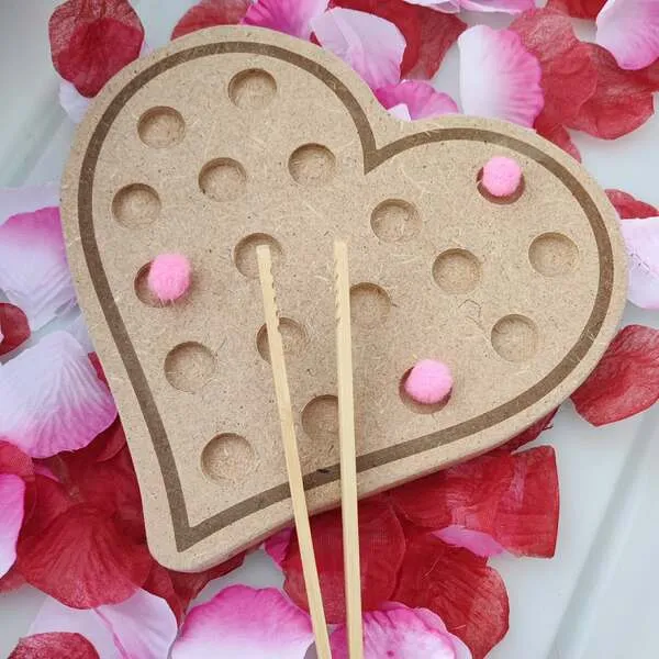 Plateau de motricité fine en bois en forme de cœur avec pompons roses et baguettes, posé sur des pétales