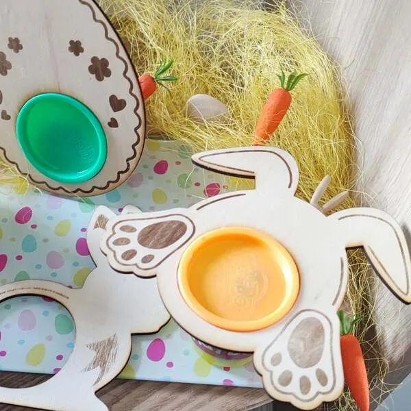Support en bois en forme de lapin avec emplacement rond pour pot de pâte à modeler, présenté sur une table sensorielle de Pâques avec œuf décoré et carottes.