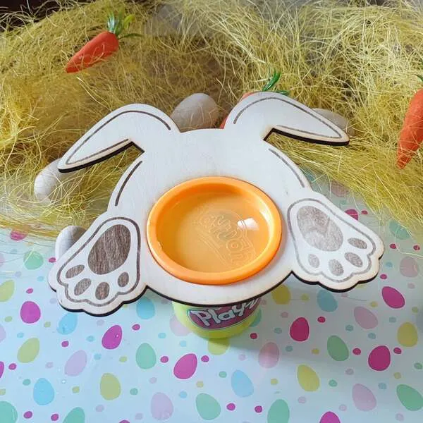 Support en bois en forme de lapin vu de dessus, avec ouverture centrale accueillant un pot de pâte à modeler, décor de Pâques avec paille et carottes.