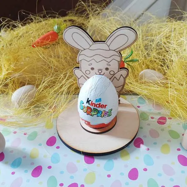 Support en bois thème Pâques avec personnage lapin sortant d’un œuf, tenant un chocolat type œuf surprise, décor paille et carottes.