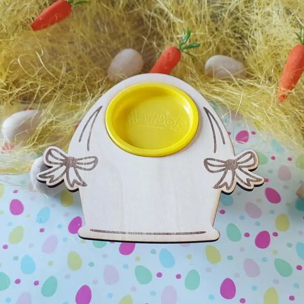 Support en bois en forme de cloche de Pâques avec ouverture centrale pour pot de pâte à modeler, décoré de nœuds gravés et présenté sur une table printanière.