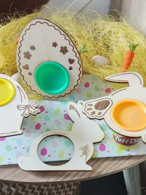 Supports en bois thème Pâques pour pots de pâte à modeler, formes œuf et lapin avec emplacements ronds colorés, posés sur une table décorée façon chasse aux œufs.